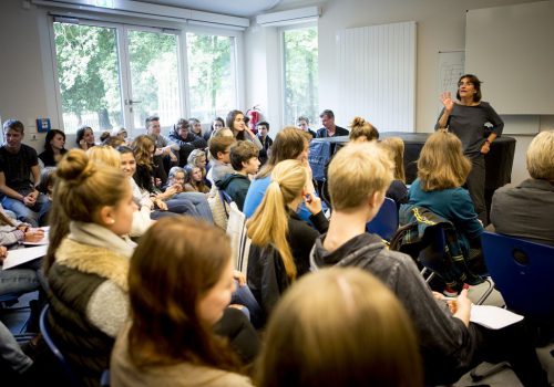 Viele Schüler sind in einem Raum und hören einer Frau zu.