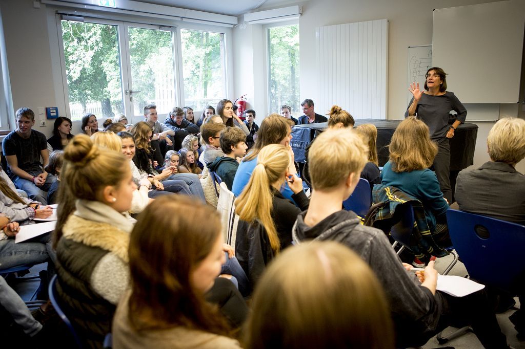 Viele Schüler sind in einem Raum und hören einer Frau zu.